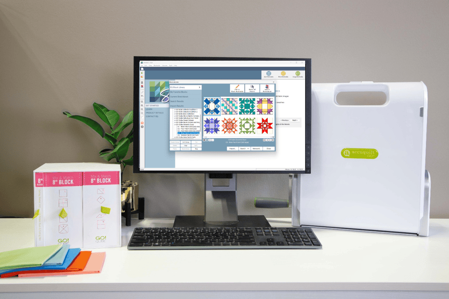 An office desk with a computer monitor displaying quilting software, a keyboard, a white fabric cutter machine, and two boxes labeled '8" Block' along with colored fabric sheets.
